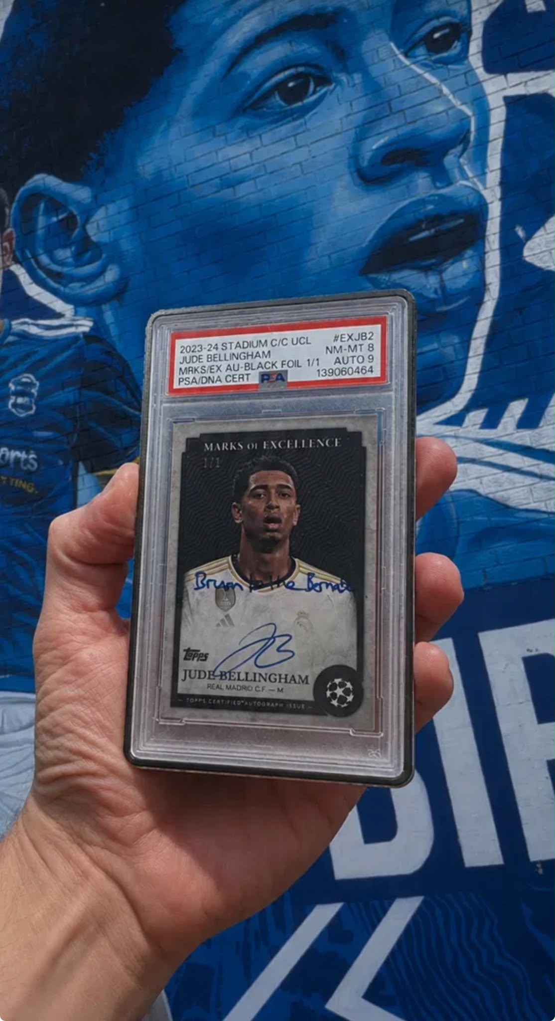 Jude Bellingham's signed one-of-one card held in front of his Birmingham mural